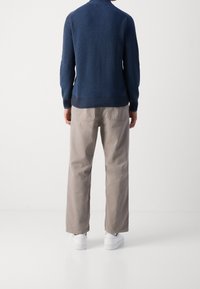 Maglione a coste blu navy con collo alto, abbinato a pantaloni beige chiaro a gamba dritta e sneaker bianche, visto da dietro.