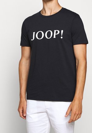 Mężczyzna w czarnej koszulce z krótkim rękawem z białym napisem „JOOP!”, w połączeniu z białymi spodniami na jednolitym jasnym tle.