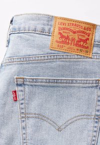 Ljusblå denimjeans med en läderetikett som har röd text och grafik. Slim fit med raka ben. Röd etikett nära fickan.