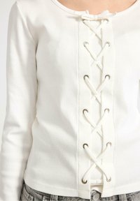 Hvid ribbet bluse med lange ærmer og snøre foran med metaløskener. Stoffet virker blødt og let tekstureret.