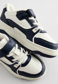Navy and white sneakers with a textured leather upper, perforated toe, and adjustable Velcro strap. Features contrasting accents and laces.