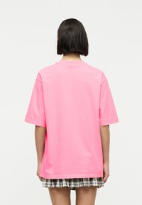 Άτομο με κοντά σκούρα μαλλιά που φοράει μια φωτεινή ροζ φαρδιά μπλούζα και μια κοντή καρό φούστα, εμφανιζόμενο από πίσω μπροστά σε λευκό φόντο.