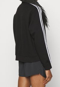 Svart sweatshirt med vita ränder längs ärmarna, tillverkad av ett mjukt tyg. Har en rund halsringning och en avslappnad passform, tillsammans med mörka shorts.