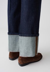 Jean en denim avec une teinture bleu foncé, ornée de détails en bleu clair au niveau des ourlets et d'une bande d'accent marron. Une chaussure en daim marron est visible.
