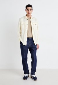 Vaalean beige -värinen puserotakki, jossa on kaksi rinta-taskua, tummansiniset farkut ja tummansiniset tennarit, joissa on keltaista korostusta. Sujuva kangas, rento istuvuus.