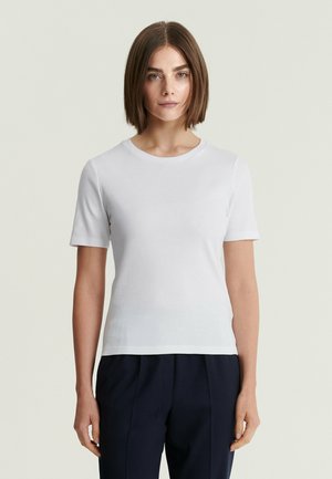 Femme aux cheveux bruns et lisses portant un t-shirt blanc uni à manches courtes et un pantalon foncé, debout devant un fond clair.