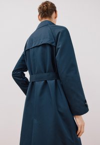 Marineblauer Trenchcoat mit einem Rückenventil, tailliertem Schnitt und auffälligem Kragen. Glatter Stoff mit strukturiertem Design und Knopfakzenten.