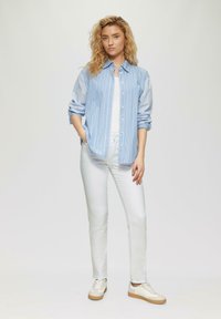 Camisa de botones a rayas azul claro y blanca, jeans blancos ajustados, zapatillas beige con acentos claros, el modelo está de pie con las manos en los bolsillos.