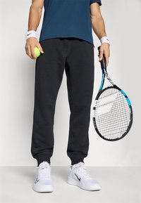 Person i sorte sweatpants, hvide sneakers og blå skjorte, som holder en tennisbold i venstre hånd og en blå-sort tennisracket i højre hånd.
