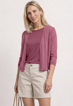Kvinde med blondt hår iført en pink cardigan og skjorte, beige shorts, holder en brun skuldertaske mod en lys væg.