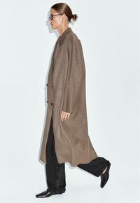 Cappotto di lana marrone con un design lungo e oversize, colletto a risvolto e doppi bottoni; abbinato a jeans neri e scarpe nere appuntite.