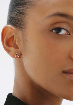 Primer plano de la oreja de una persona con el cabello liso recogido hacia atrás, que lleva un pequeño pendiente plateado angular con piedras incrustadas.