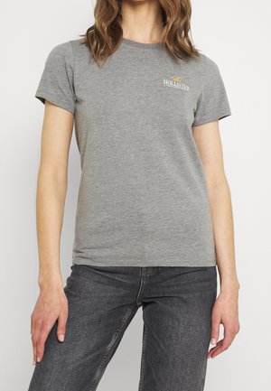 Frau trägt ein graues Hollister-T-Shirt und dunkelgraue Jeans, steht mit locker an den Seiten hängenden Armen vor einem einfarbigen Hintergrund.