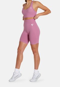 Kobieta poprawiająca różowe, wysokie szorty sportowe oraz dopasowany stanik sportowy z plecakiem, w białych skarpetkach i butach do biegania Nike.
