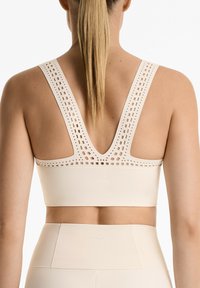 Light beige sports bra with wide straps featuring cut-out patterns, a deep V-back, and a smooth, seamless design. Prominent "OYSHO" logo.