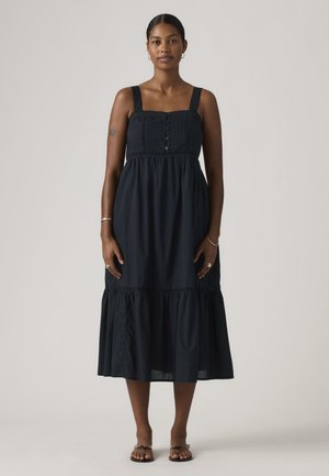 Femme debout portant une robe midi noire sans manches avec des boutons sur la poitrine et des sandales noires sur un fond uni.