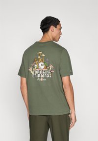 Πράσινο ελιάς βαμβακερό t-shirt με πολύχρωμο γραφικό μανιταριού και κείμενο «FORAGING FAIRWAYS» στην πλάτη. Κοντά μανίκια και στρογγυλή λαιμόκοψη.