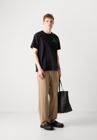 Svart bomull T-skjorte med grønn trekantgrafikk, kombinert med beige bukser. Modellen holder en svart tote bag og har på seg svarte sko.