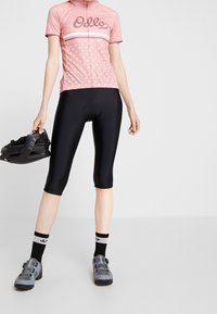 Różowa koszulka rowerowa w białe kropki z logo, w zestawieniu z czarnymi szortami rowerowymi. Model trzyma czarną butę rowerową.