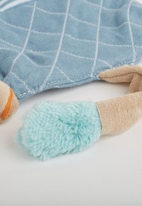 Couverture en tissu bleu doux avec un motif de grille cousu blanc et des parties de jouets en peluche texturées beige et bleu clair attachées sur une surface blanche.
