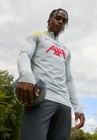 Harmaa pitkähihaisella jalkapalloharjoitustopilla, jossa on keltaisia yksityiskohtia ja punainen 'AXA'-logo, yhdistetty tummanharmaisiin housuihin, joissa on punainen 'L.F.C.'-embleemi. Pelaaja pitää jalkapalloa.