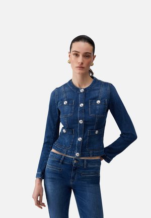 Frau trägt eine taillierte blaue Jeansjacke mit silbernen Knöpfen und passende blaue Jeans, steht mit einer Hand in der Hüfte vor weißem Hintergrund.