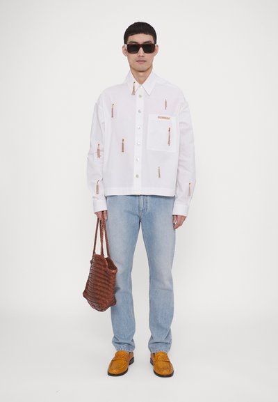 Hvid skjorte med orange broderi, lyseblå jeans, brun vævet taske og gule teksturerede sko. Solbriller fuldender looket.