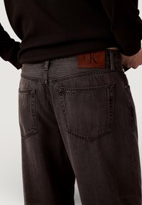 Jean en denim gris foncé avec un gros plan sur les poches arrière et une étiquette en cuir marron Calvin Klein. Présente une coupe classique et une texture subtile.