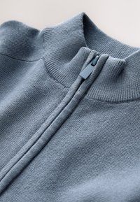 Maglione azzurro chiaro a maglia con colletto alto e chiusura con zip frontale. Presenta una texture a coste e cuciture lisce e uniformi in tutto il capo.