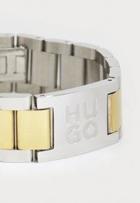 Metallarmbånd med vekslende sølv- og gullkjeder. Har en fremtredende sølvlås gravert med "HUGO." jevn tekstur og slank design.