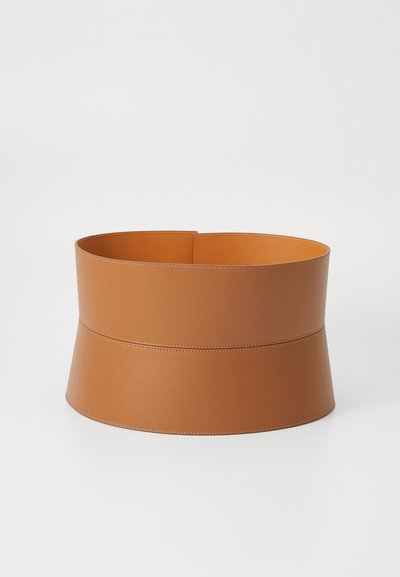 Panier rond en cuir de couleur tan avec une texture lisse, présentant un large dessus et une base plus étroite, accentué par une couture horizontale autour du centre.