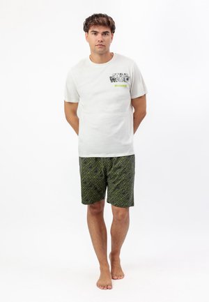 Joven de pie descalzo vestido con una camiseta blanca de Star Wars y pantalones cortos negros con un patrón de texto amarillo de Star Wars sobre fondo blanco.