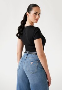 Femme aux longs cheveux noirs en queue de cheval, portant un haut noir ajusté à manches courtes et un jean bleu taille haute avec un logo de marque visible sur la poche.