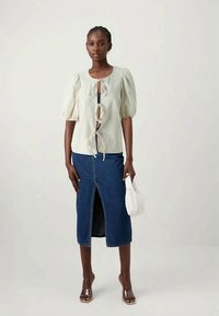 Μπλούζα με φουσκωτά μανίκια και κορδόνια στον λαιμό, συνδυασμένη με φούστα από μπλε τζίν μέσου μήκους με σχισμή μπροστά και λευκή τσάντα.