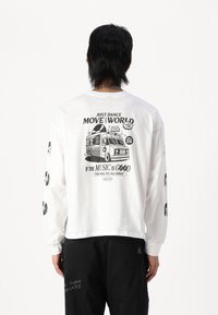 White long-sleeve t-shirt featuring printed graphics on the back and sleeves, including a record shop design and text in black. Black pants.