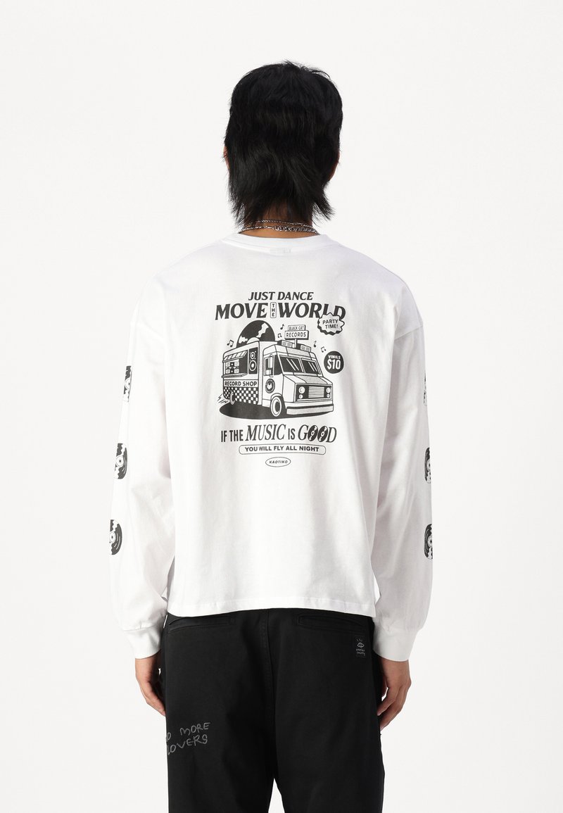 White long-sleeve t-shirt featuring printed graphics on the back and sleeves, including a record shop design and text in black. Black pants.