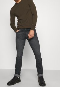 Hombre con camiseta de manga larga color verde oliva, jeans ajustados gris oscuro remangados, zapatillas negras, de pie contra un fondo liso de color gris claro.