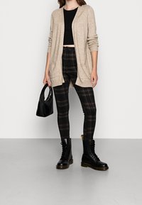 Donna che indossa un cardigan beige, un crop top nero, pantaloni a quadretti e stivali di combattimento neri, mentre tiene una piccola borsa nera su uno sfondo semplice.