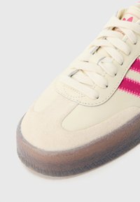 Krēmkrāsas un rozā sporta apavi, izgatavoti no zamšāda un ādas materiāliem, ar apaļu purngalu, biezām caurspīdīgām zolēm un plakaniem kokvilnas auklām.