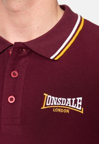 Burgundijos spalvos polo marškiniai su tekstūruotu audiniu, trimis mygtukais, ir kontrastingu dryžuotu apykakle. Papuošti geltonu ir baltu siuvinėjimu.