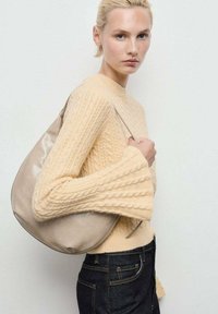 Donna bionda che indossa un maglione beige a treccia e jeans scuri, con una borsa in pelle beige a tracolla su uno sfondo semplice.