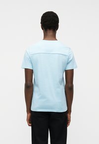 Άνδρας που φοράει γαλάζιο κοντομάνικο πουκάμισο και μαύρο παντελόνι, στέκεται γυρισμένος από την πλάτη μπροστά σε απλό λευκό φόντο.