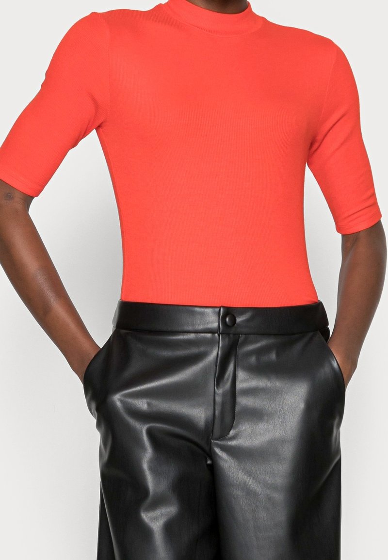 Osoba w dopasowanym, jaskrawym pomarańczowym topie z krótkim rękawem, włożonym do wysokich, czarnych skórzanych spodni, z rękami w kieszeniach.