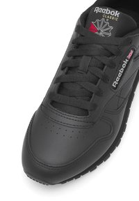 Fekete bőr sportcipő kerek orral, lapos fűzőkkel, finom perforációkkal és fehér, valamint piros Reebok márkajelzéssel a nyelvén és az oldalán.