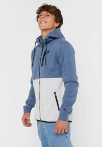 Giovane che indossa una giacca con cappuccio blu e grigia chiara con dettagli neri della zip, sorridente e con lo sguardo rivolto verso l'alto.
