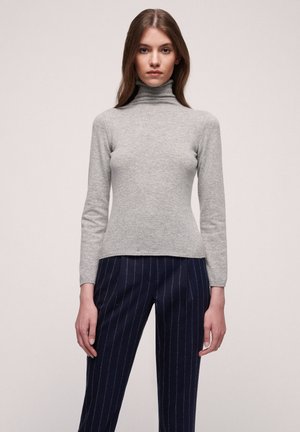 Grauer Rollkragenpullover aus weichem, gestricktem Stoff mit langen Ärmeln. Kombiniert mit dunkelblauen Hosen mit Nadelstreifen, was eine figurbetonte Silhouette ergibt.