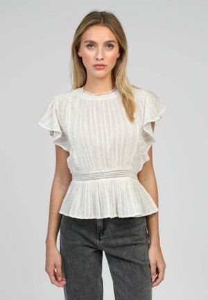 Femme portant un chemisier blanc texturé avec des manches à volants et une taille peplum, associé à un jean taille haute gris foncé.