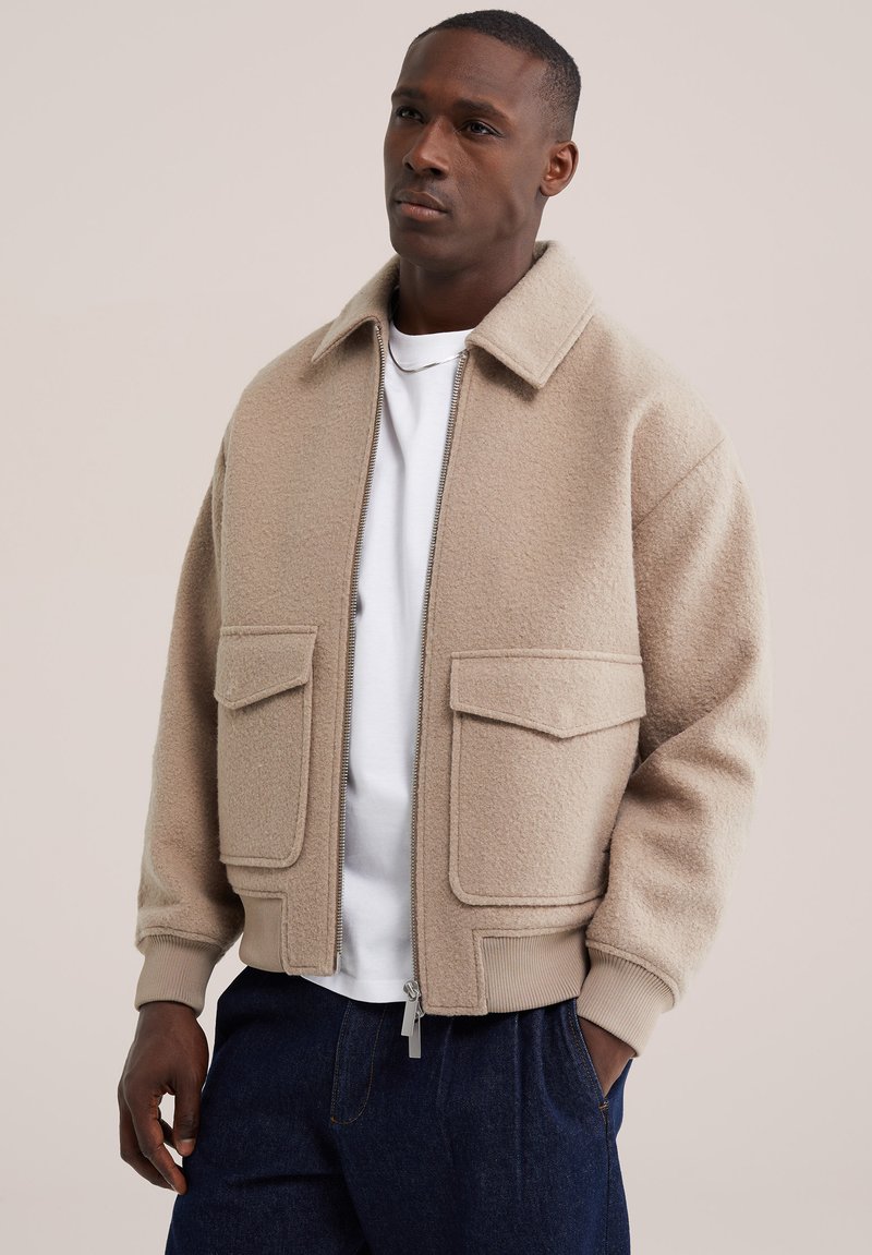 Homme portant une veste en laine beige zippée avec de grandes poches frontales sur une chemise blanche et un pantalon bleu foncé, sur fond uni.