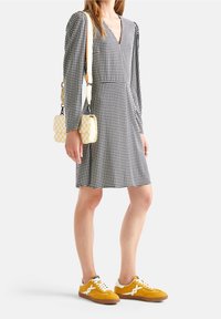 Abito a righe bianche e nere con maniche lunghe, scollo a V profondo, abbinato a una borsa a quadri gialla e sneakers senape con accenti bianchi.