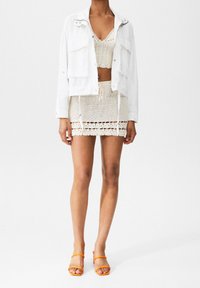 Biała kurtka dżinsowa na kremowym komplecie z szydełkowanej bluzki i spódnicy, z detalami z otwartym splotem i wiązaniem w talii. Do tego pomarańczowe sandały.
